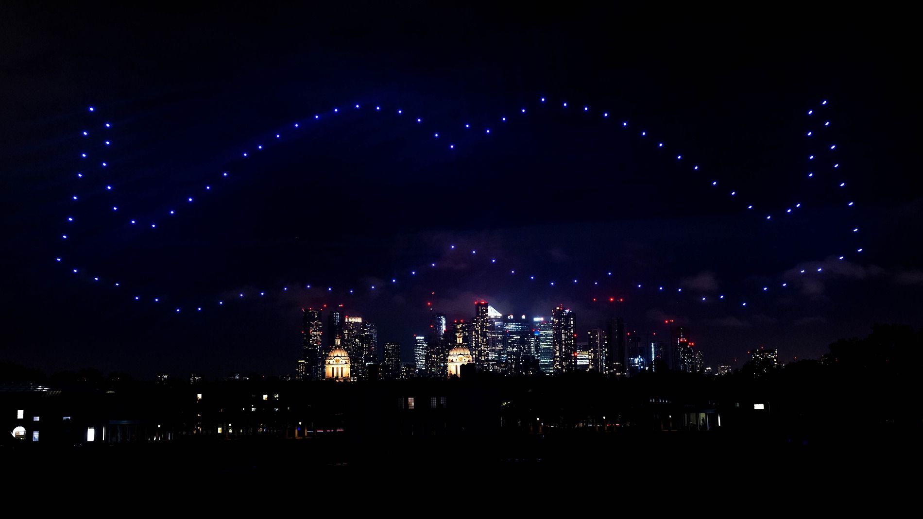 Photo of illuminated flying drones, forming a moustache shape in the night sky.