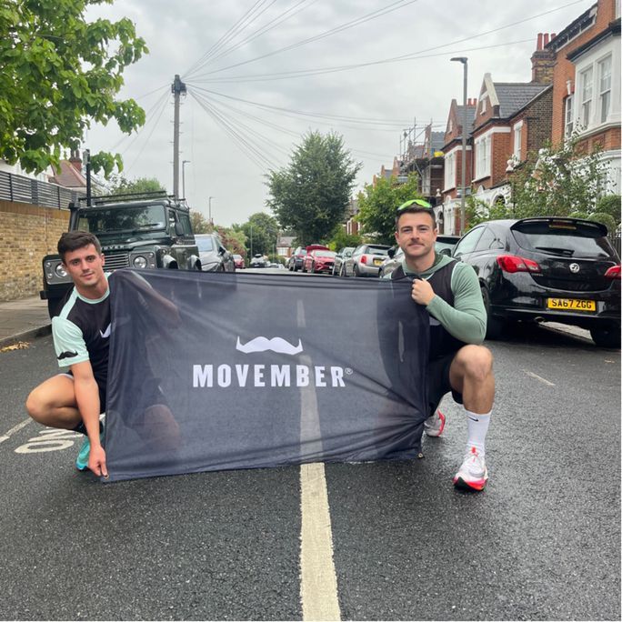 Photo of two athletic men wearing activewear, holding a Movember-branded flag to camera.