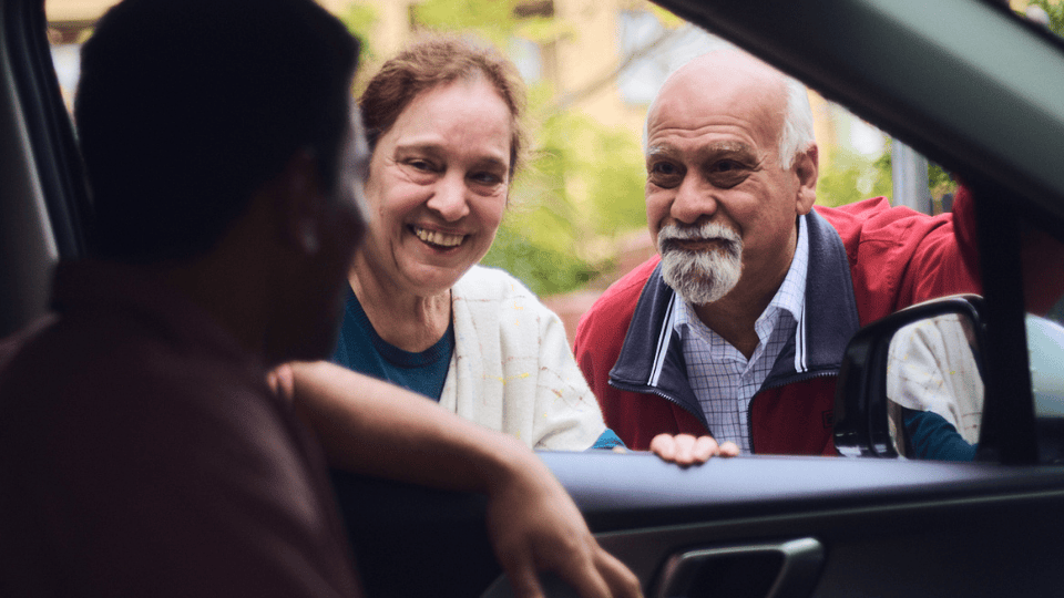 A middle-aged man in a stationary car, talking to his senior mother and father through the open driver-side window.