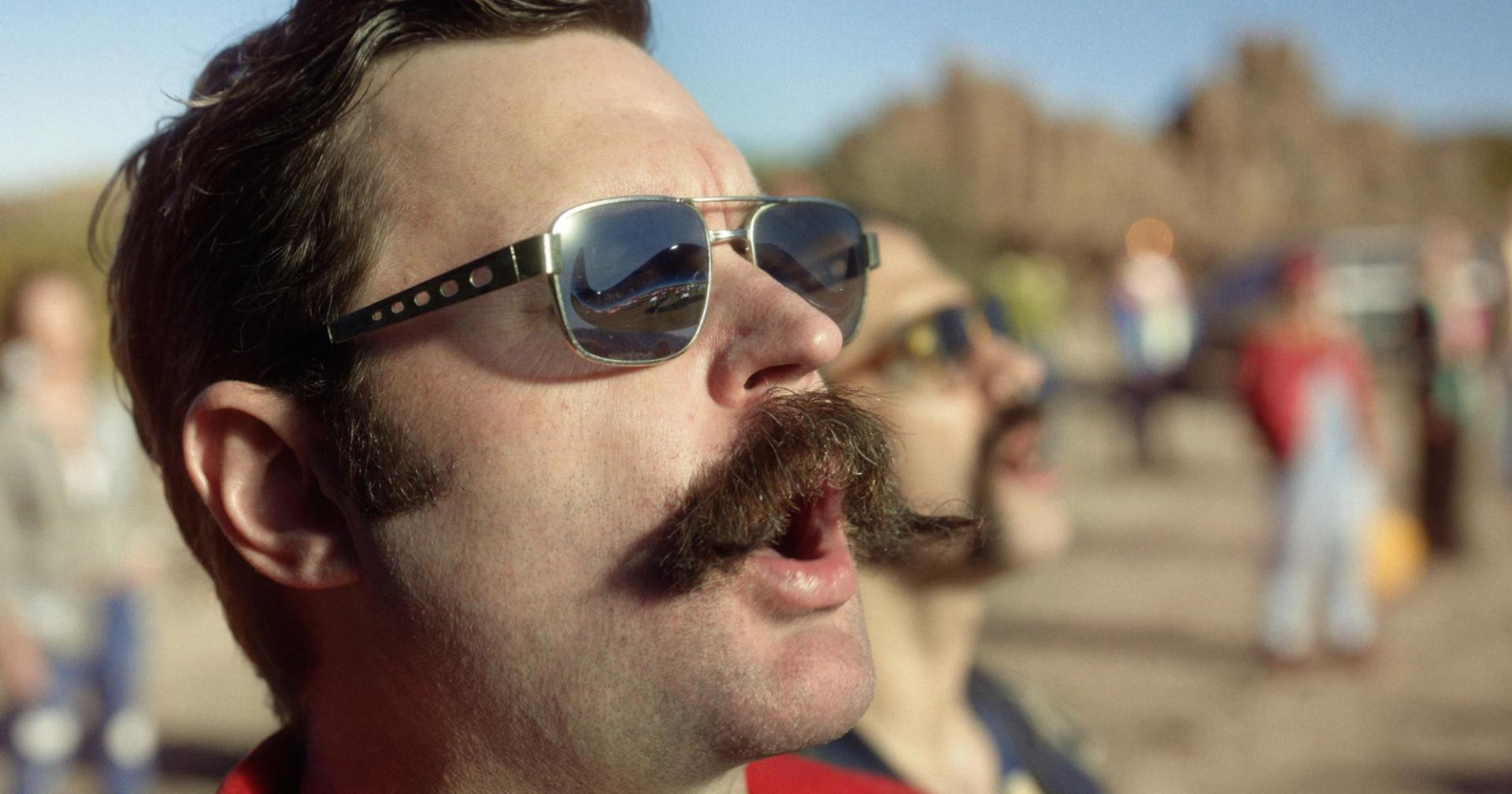 Photo of a man with a magnificent moustache, calling out, near a group of people.